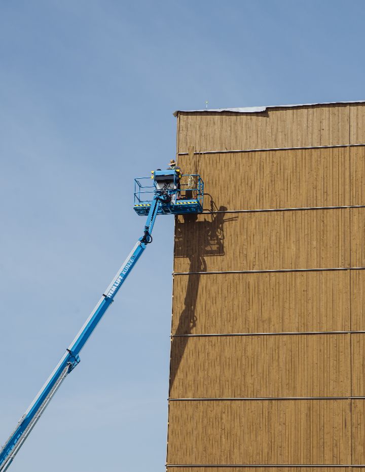 Arbeiter auf Hebebühne montieren vertikale Holzverkleidung an einer hohen Fassade