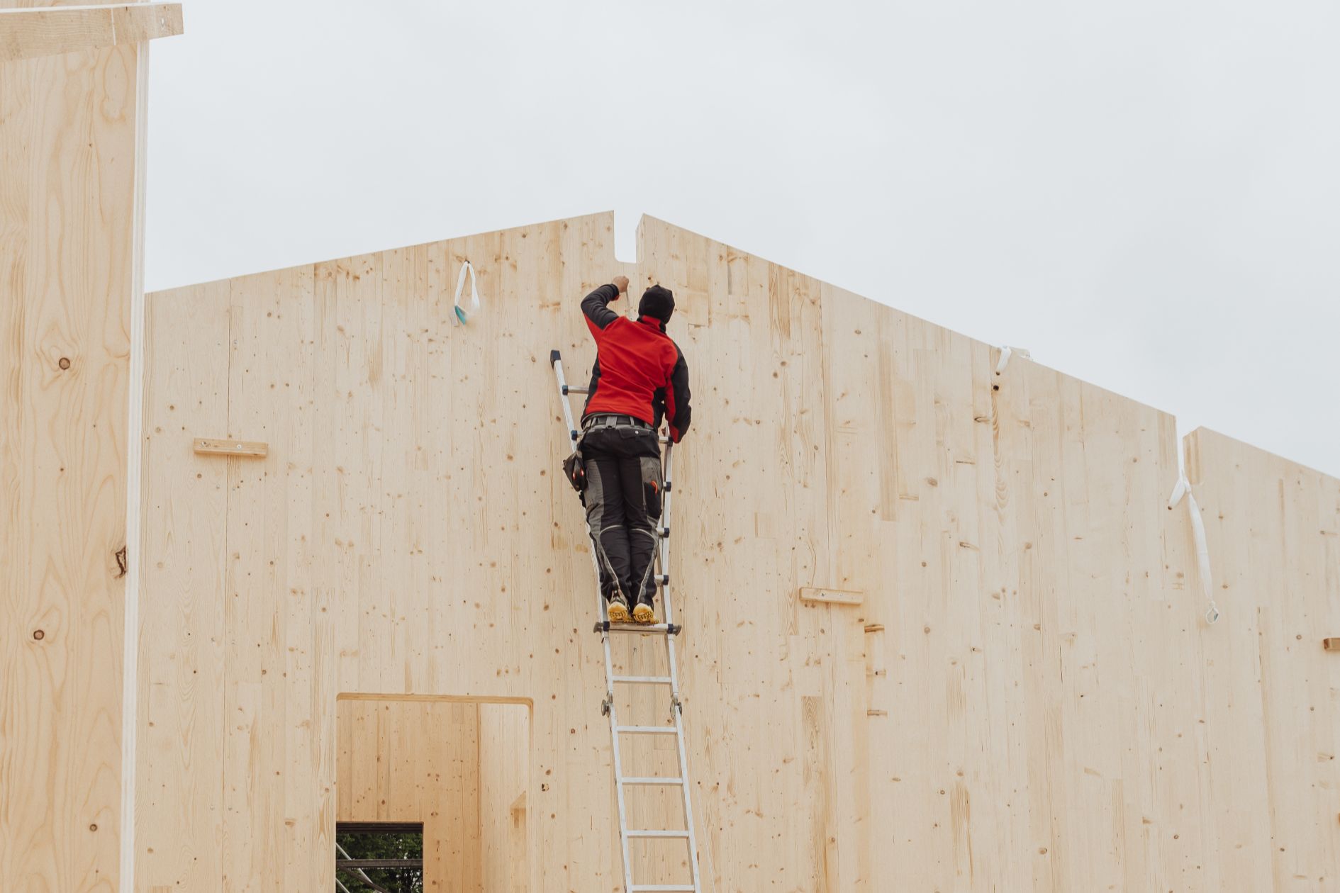Handwerker auf Leiter arbeitet an der Holzfassade eines Gewerbebaus.