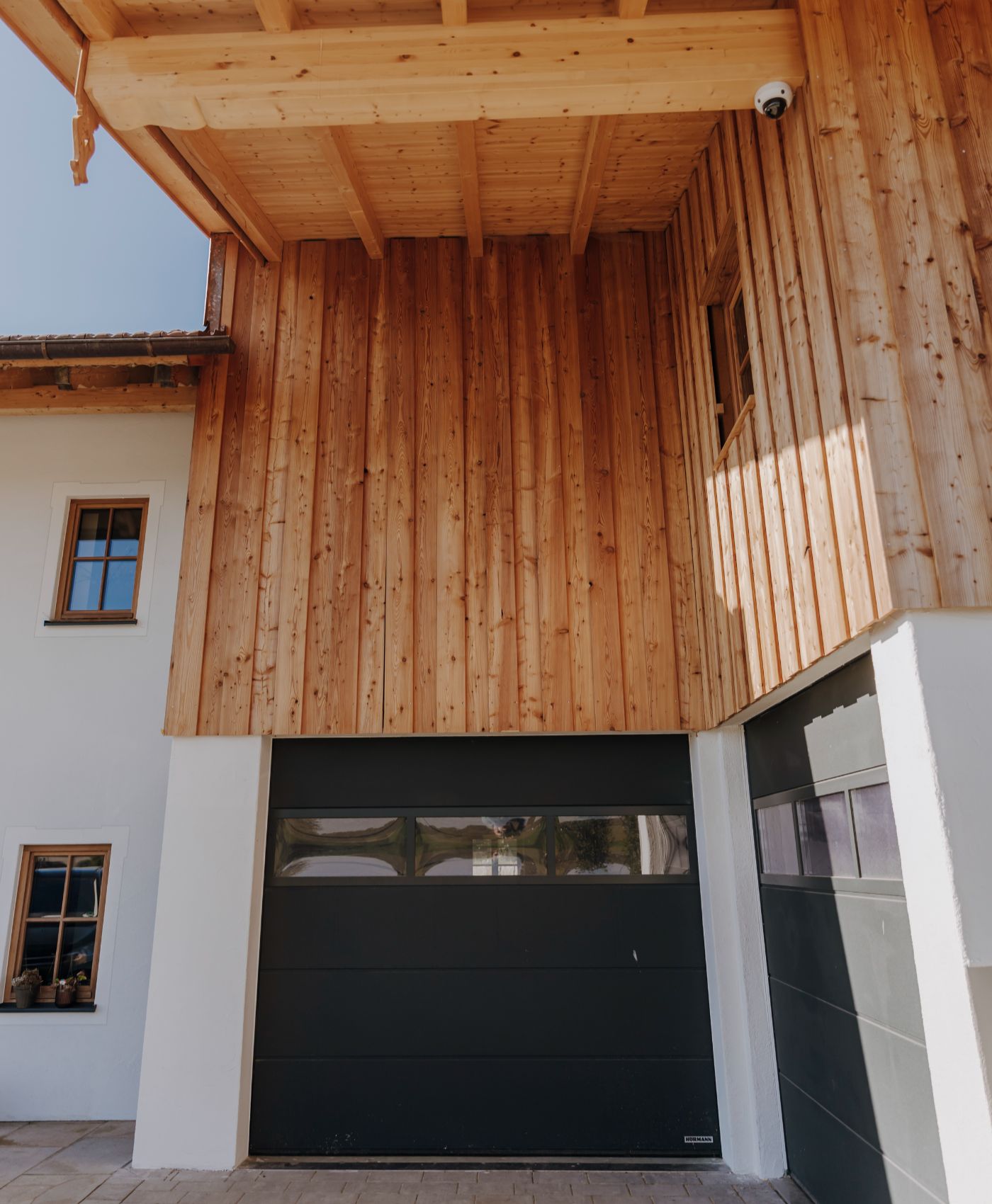 Hausfassade mit Holzverkleidung, zwei Garagentoren und Fenstern mit Holzrahmen.