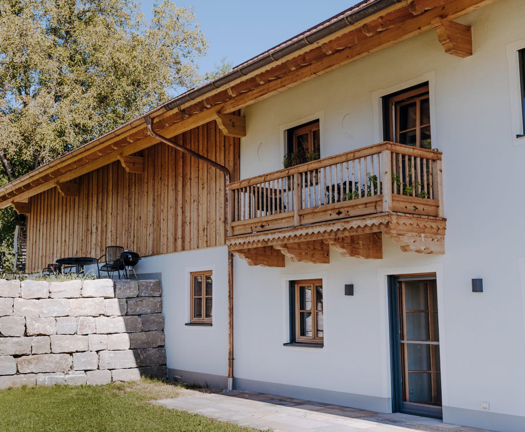 Wohnhaus mit Holzfassade, Balkon aus Holz, weißen Wänden und angrenzender Terrasse mit Steinmauer.