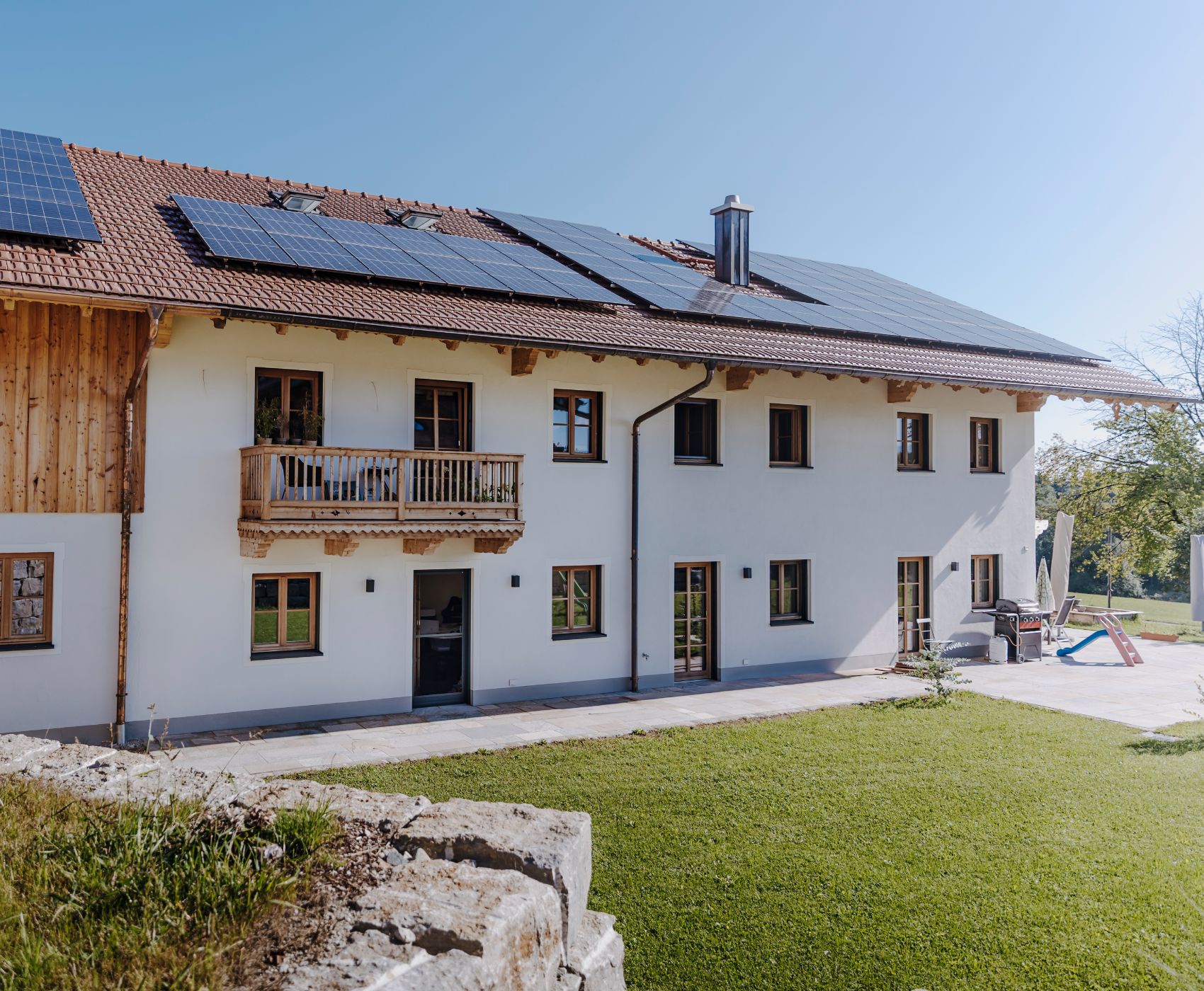 Großes Wohnhaus mit Holz- und Putzfassade, Balkon, Solarpaneelen auf dem Dach und angrenzendem Garten.