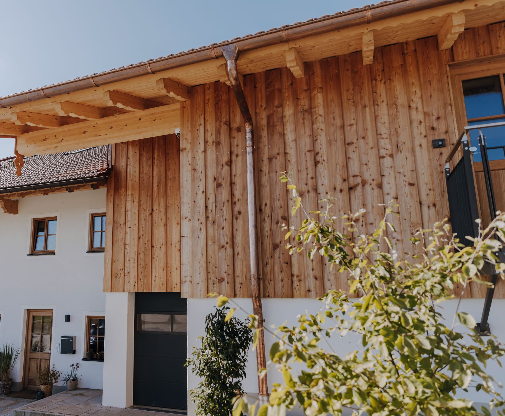 Hausfassade mit Holzverkleidung, Kupferregenrinne, schwarzem Garagentor und Außentreppe neben grünen Pflanzen.
