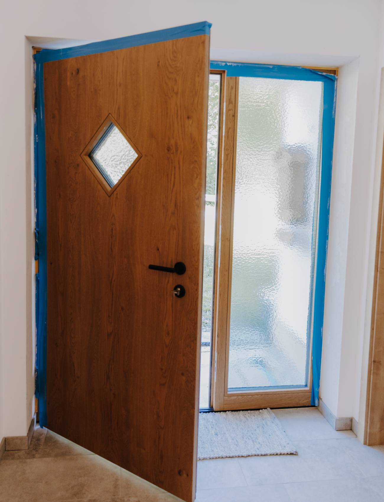 Geöffnete Holztür mit rautenförmigem Fenster, daneben Glas-Seitenteil mit Milchglas im Eingangsbereich