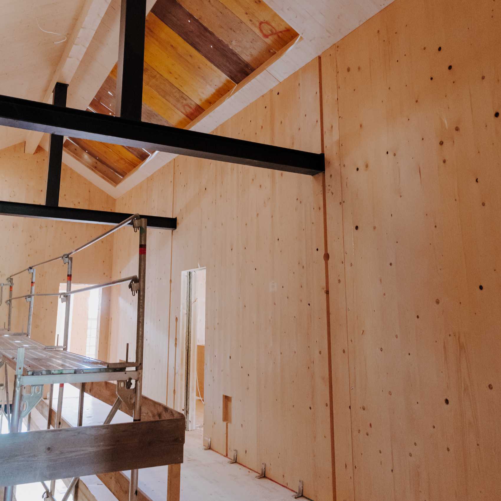 Innenansicht eines Neubaus mit Holzverkleidung, schwarzem Stahlträger und Gerüst, Decke mit Dachfensteröffnungen.