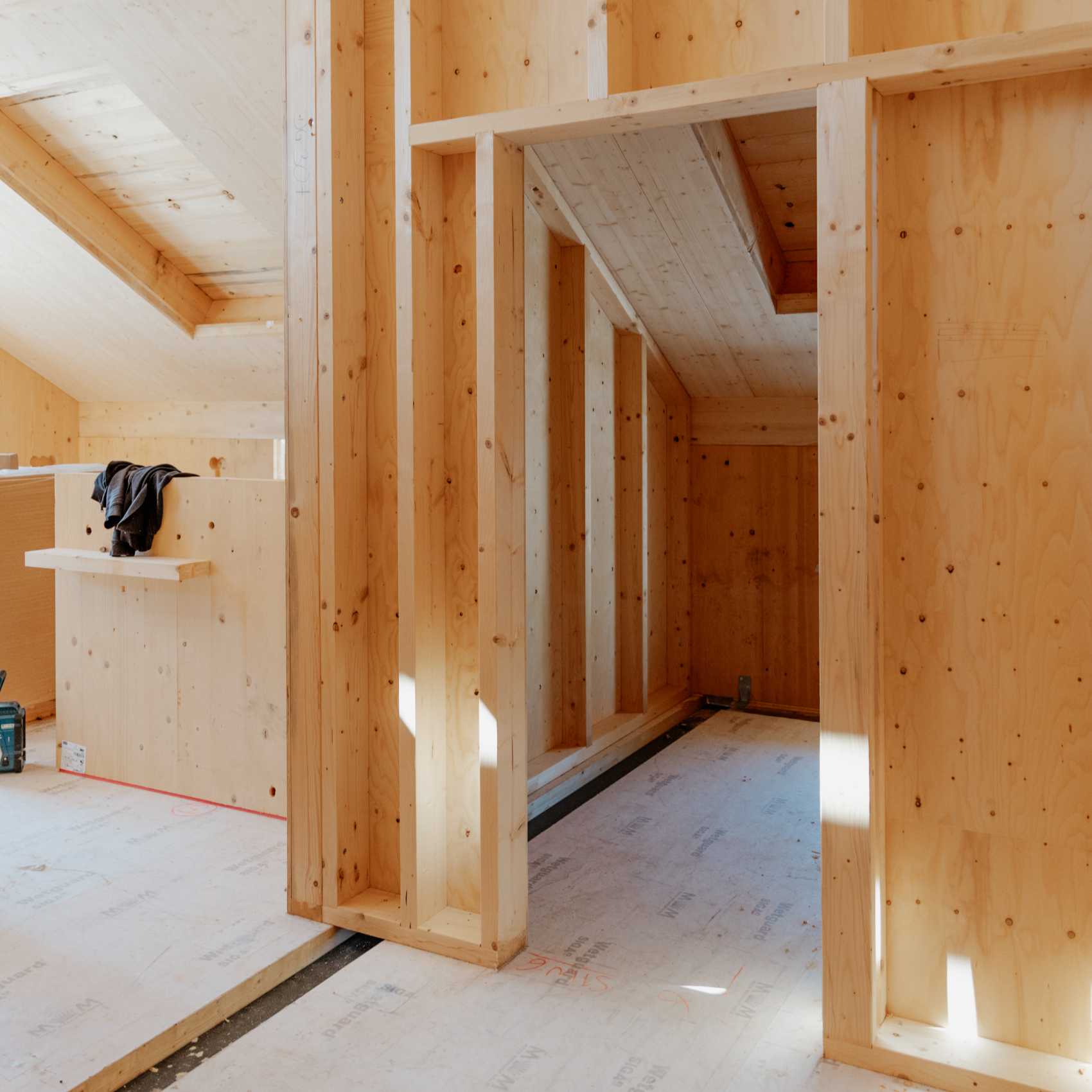 Innenraum eines Neubaus mit Holzwänden und sichtbaren Trennwänden, teilweise verkleideter Dachschräge und offenem Bodenbereich.
