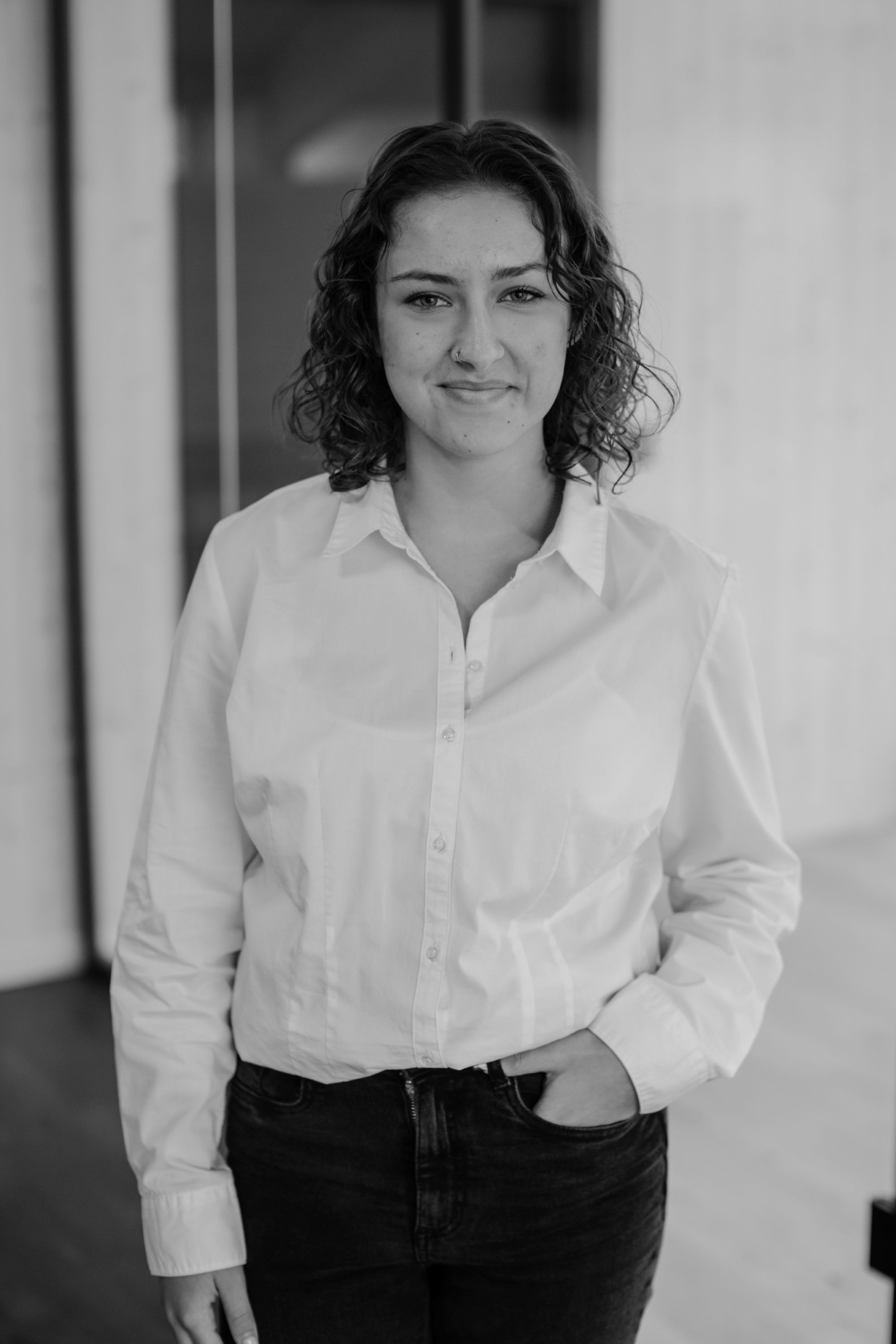 Lea Rasch trägt weiße Bluse und dunkle Hose, steht mit einer Hand in der Hosentasche vor einer Glaswand.