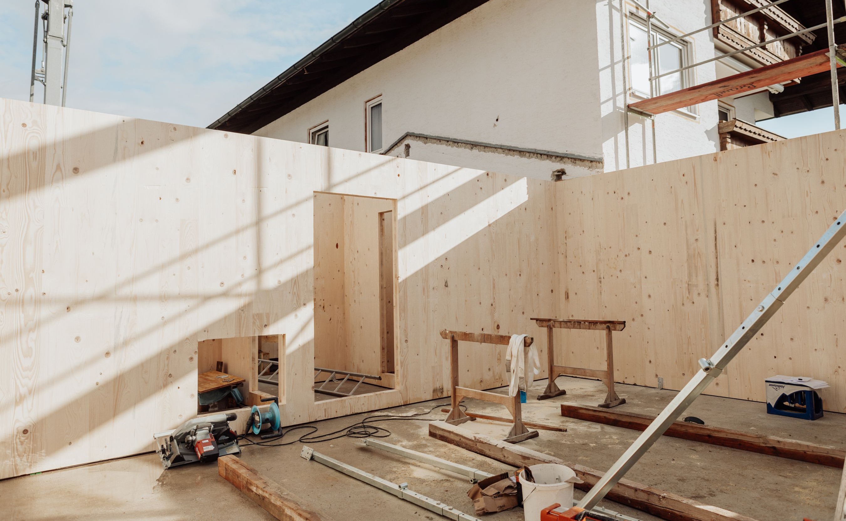 Montierte Brettsperrholzwände im Anbau an ein Bestandsgebäude, vorbereitete Wandöffnungen und Baustelleneinrichtung im Rohbauzustand.