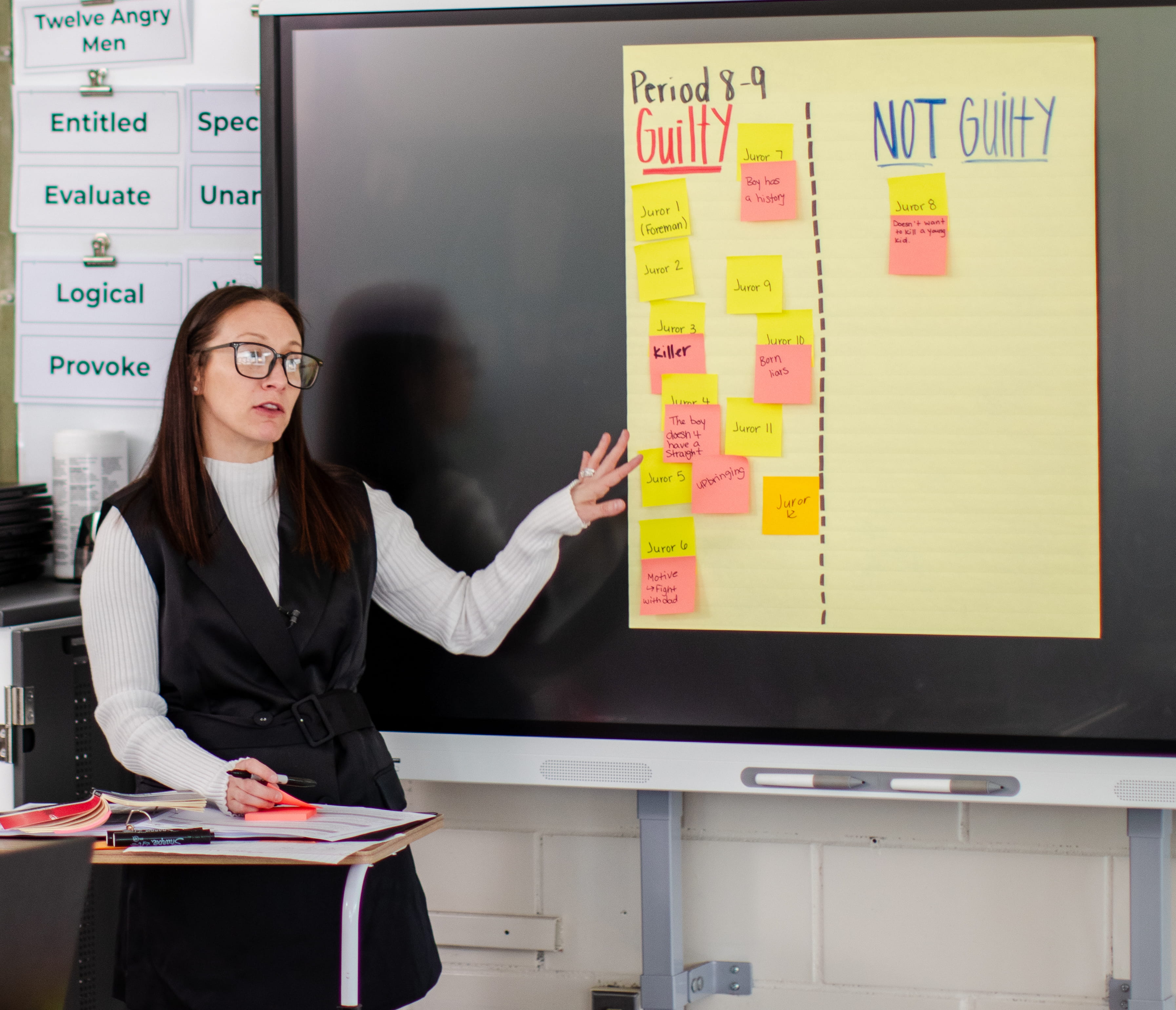 Teacher in glasses presenting a chart with sticky notes labeling jurors as Guilty or Not Guilty during a classroom activity.