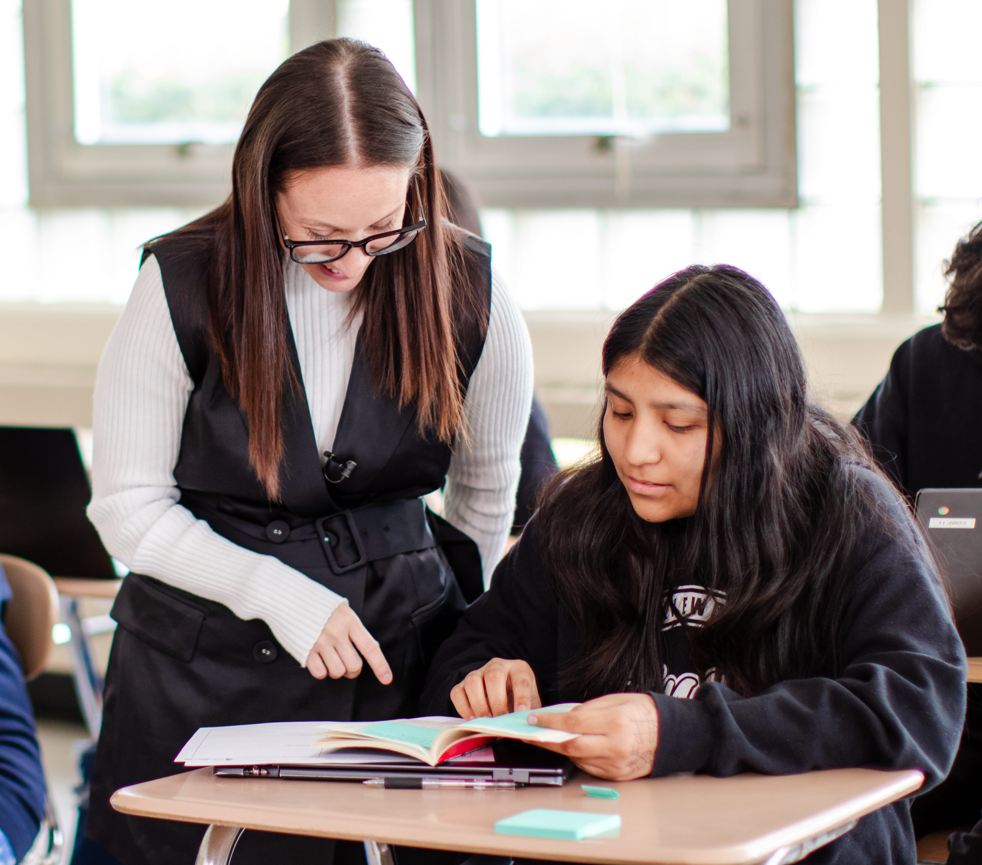 Teacher with glasses helping a student who is reading a book at a classroom desk.