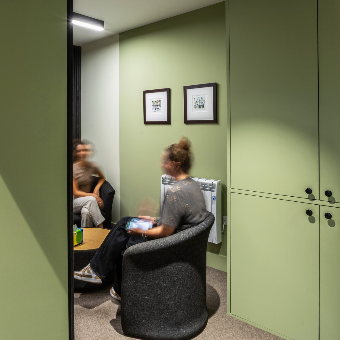 Small therapy room with green walls, two people talking in dark chairs, and soft, calming finishes.