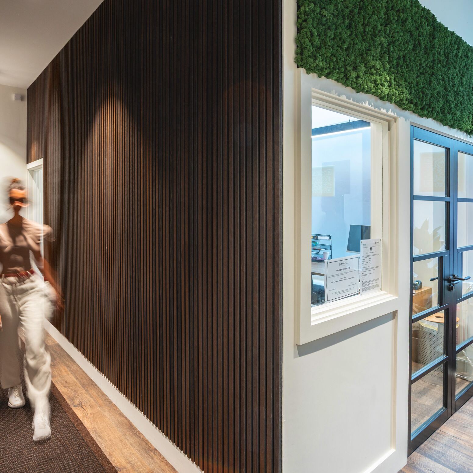 Hallway with timber slat walls, moss detail above a window, and a person walking through the calm, modern space.