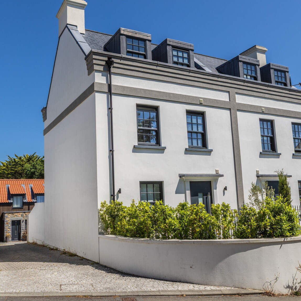 Elegant three-storey white townhouse with grey trim and dormer windows, set against a blue sky and framed by lush greenery