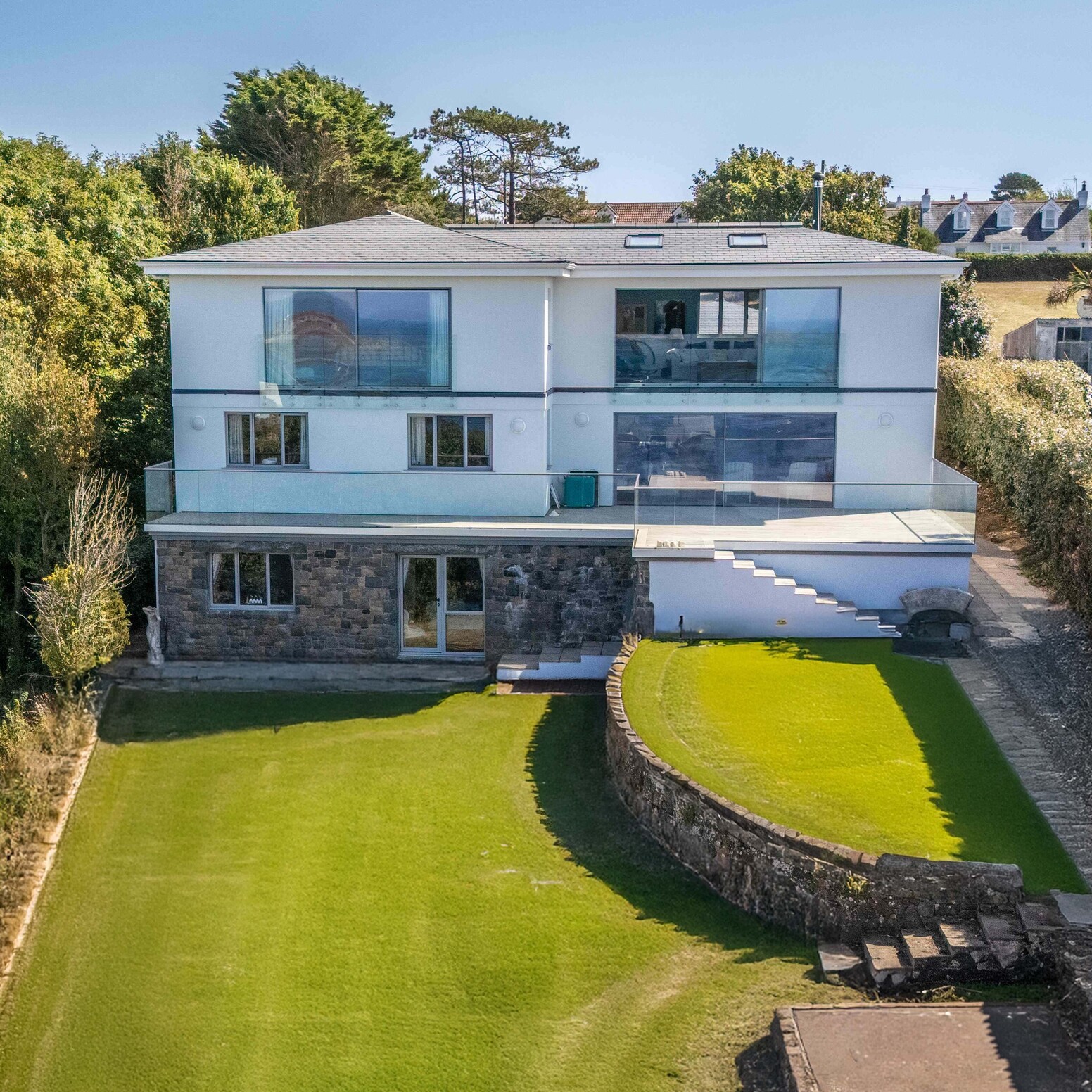 Rear elevation of a two-storey white house. Glass balconies, stone lower ground, terraced garden and sea views reflected in t