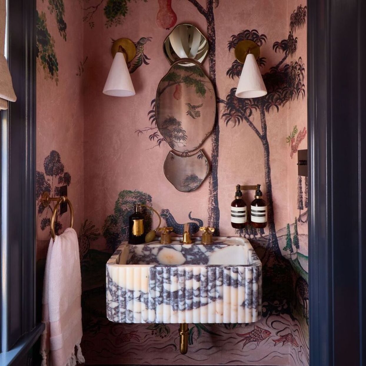 Bold small bathroom with pink scenic wallpaper, marble sink, brass taps and wall lights for a luxe boutique cloakroom look.