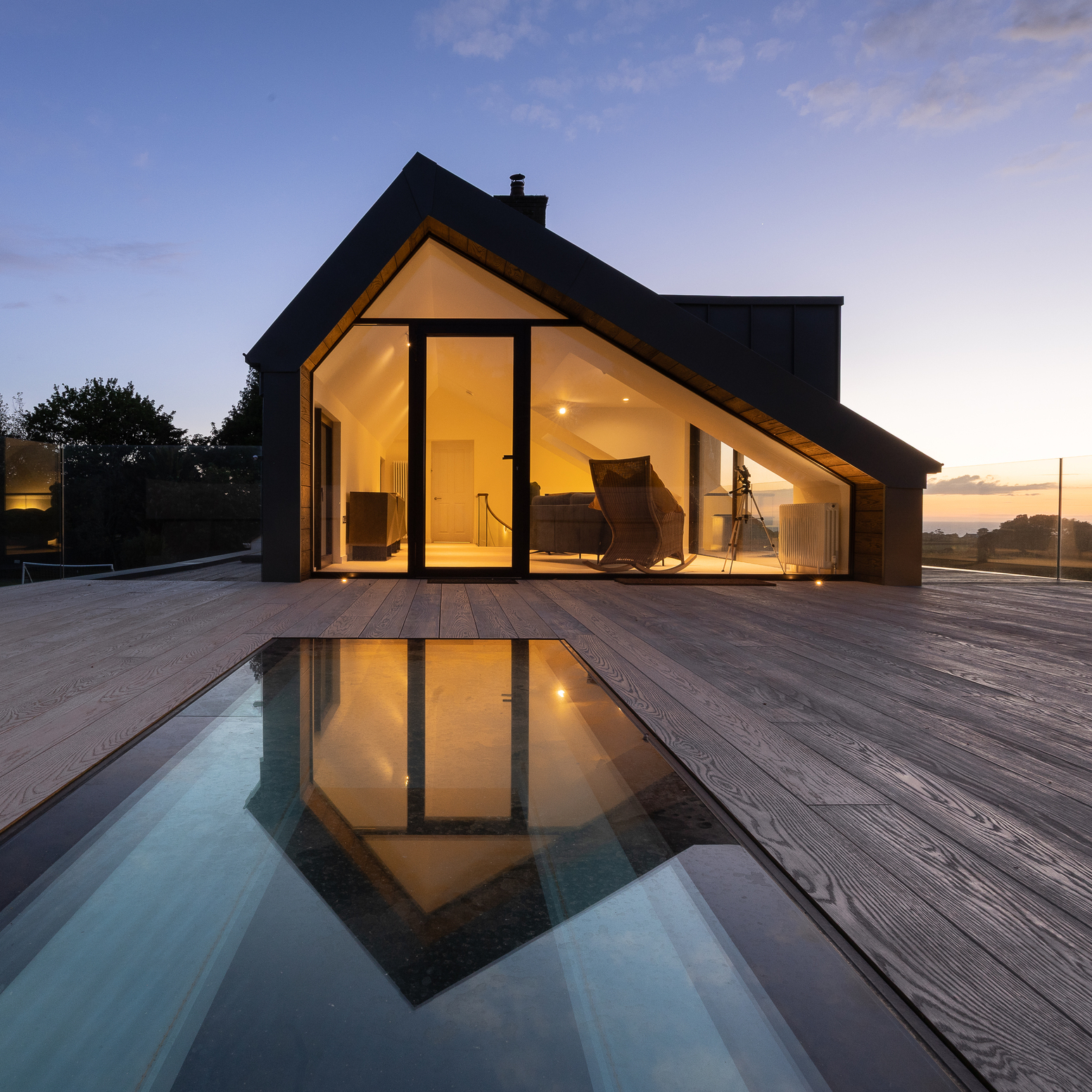Modern coastal home at dusk with large glass doors, timber decking and sea views.