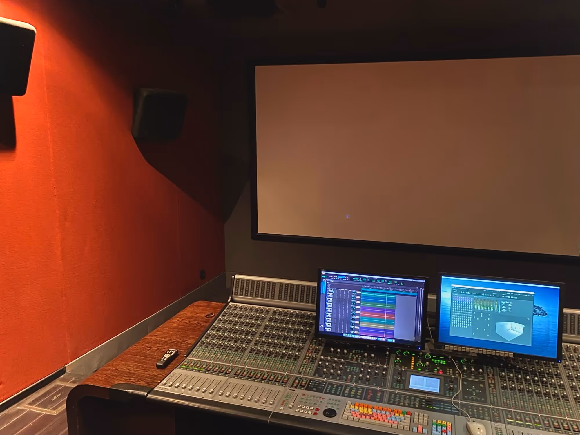 Wide-angle view of a Dolby Atmos studio featuring a professional mixing console, dual monitors with a digital audio workstation (DAW) session, a large projection screen, and mounted surround sound speakers, highlighting an immersive environment for advanced audio production.