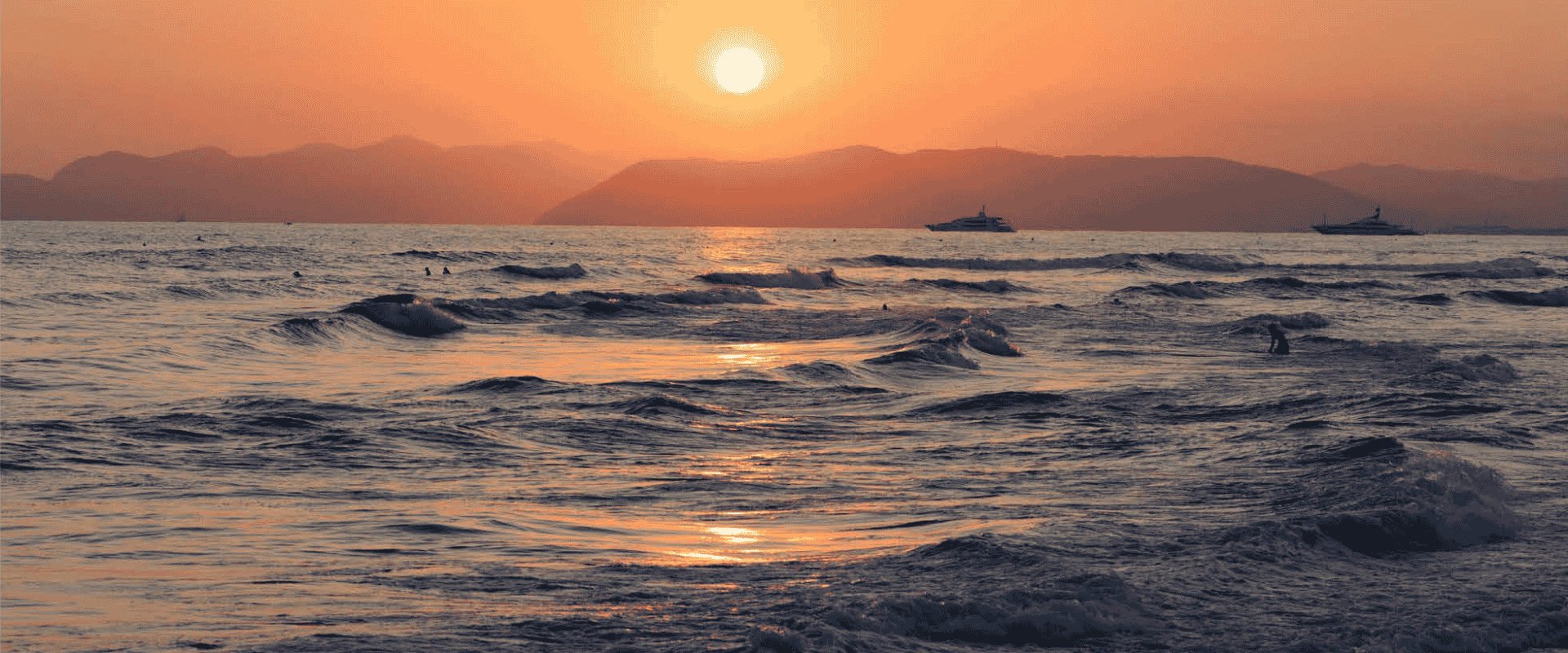 Sun setting over the sea with soft waves, distant hills, and boats in the background.
