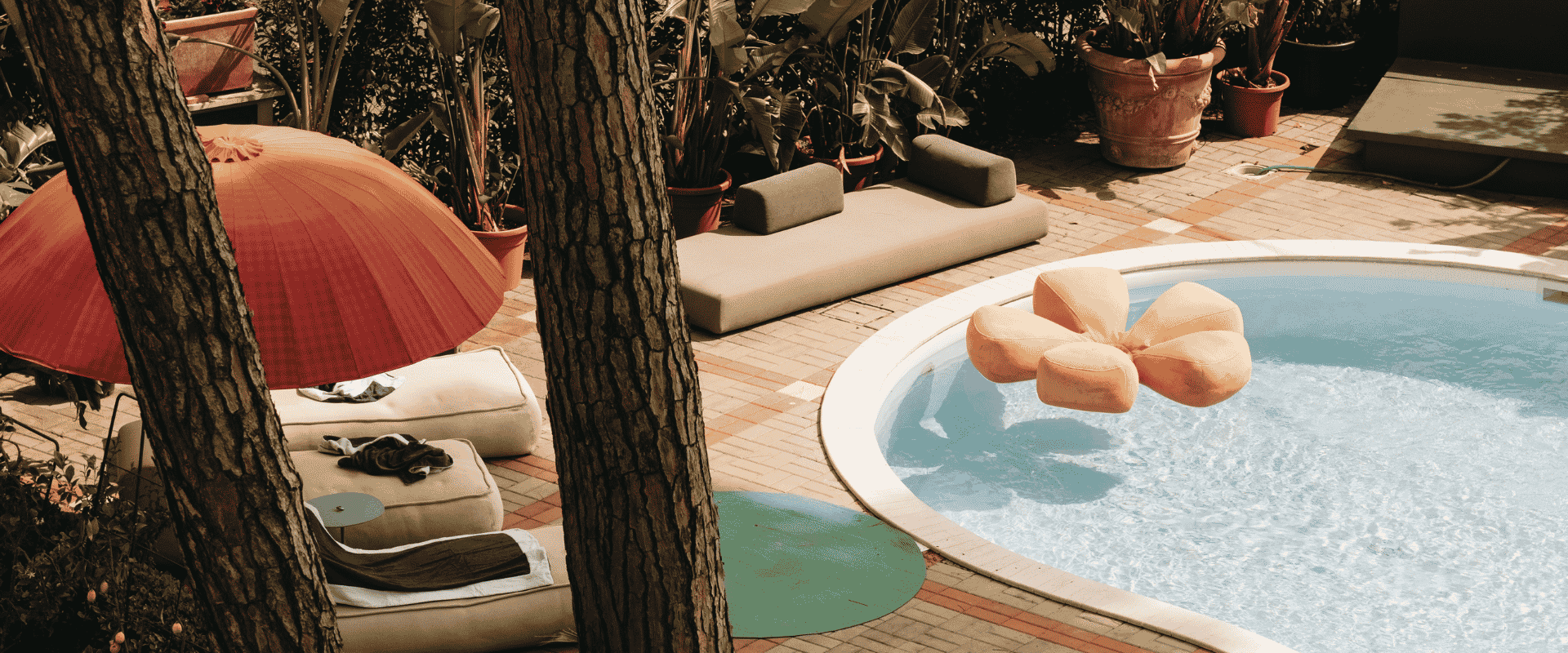 Patio soleggiato a bordo piscina con ombrellone rosso, materassi a terra e piante tropicali.