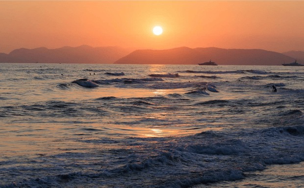 Golden sunset over the ocean with gentle waves, silhouettes of swimmers, and distant mountains.