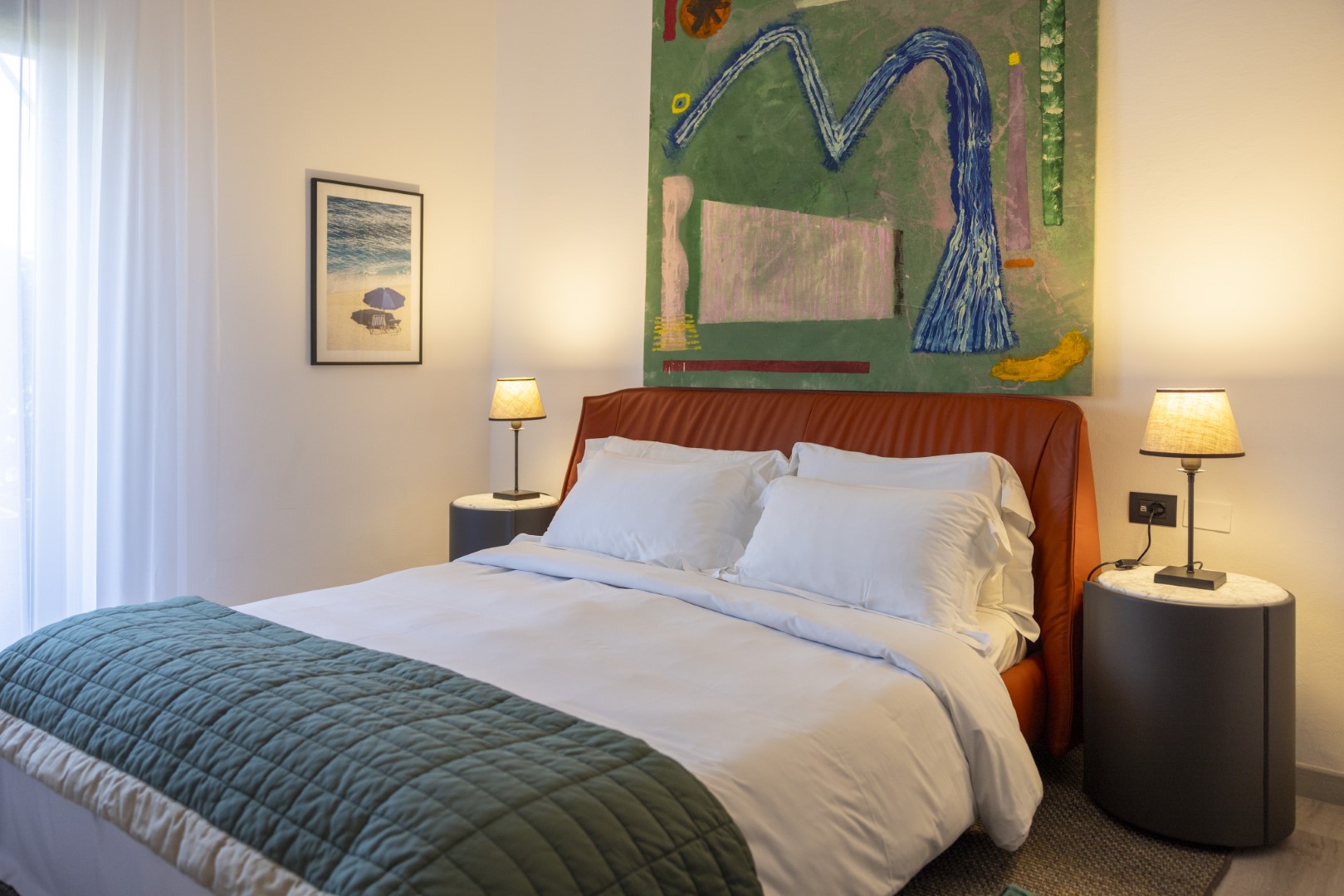 Bedside scene with a neatly made bed, white pillows and sheets, a wooden nightstand, and a lit table lamp with a warm orange shade next to rust-colored curtains.