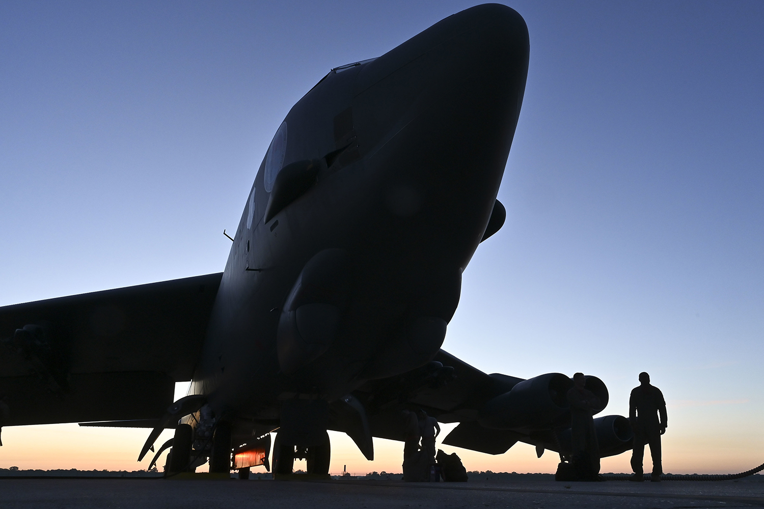 Silhouetted military aircraft on a runway at sunset with three people standing and sitting near its landing gear.