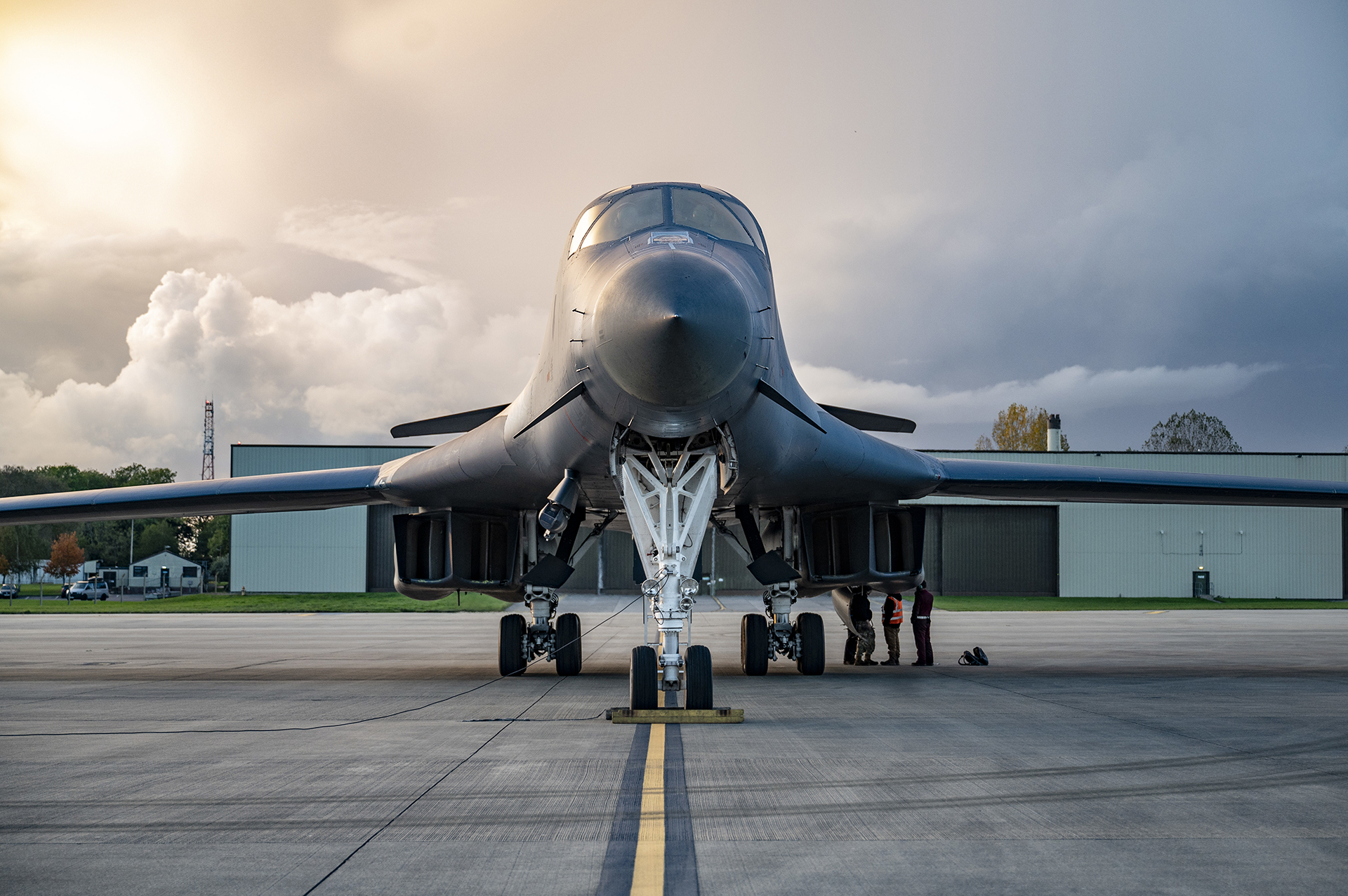 Front view of a large military bomber aircraft on a runway with three people standing near its landing gear and hangars in the background under a cloudy sky.