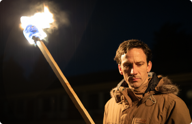 Man with gray hair and beard in navy suit and tie sitting with hands clasped, looking thoughtful.