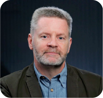 Smiling woman with long blonde wavy hair wearing a dark blazer and white blouse against a textured gray background.