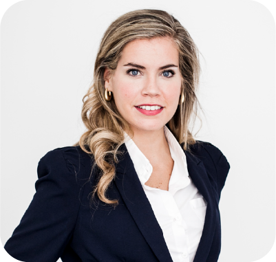 Smiling man with short brown hair wearing a dark jacket and blue shirt outdoors with blurred green foliage background.