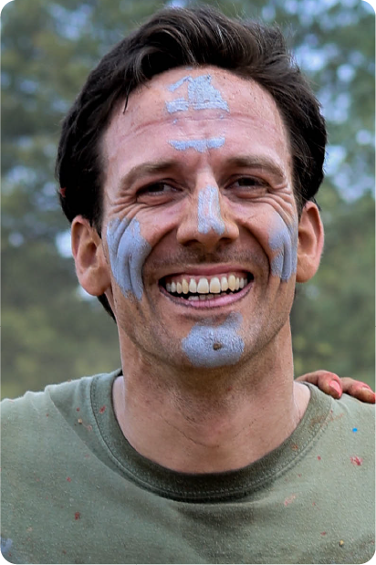Man with grey beard and curly hair dressed in a dark suit and tie, sitting with fingers interlaced in front of a dark background.