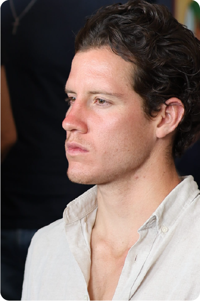 Man with grey beard and curly hair dressed in a dark suit and tie, sitting with fingers interlaced in front of a dark background.