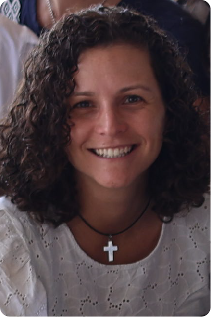 Man with grey beard and curly hair dressed in a dark suit and tie, sitting with fingers interlaced in front of a dark background.