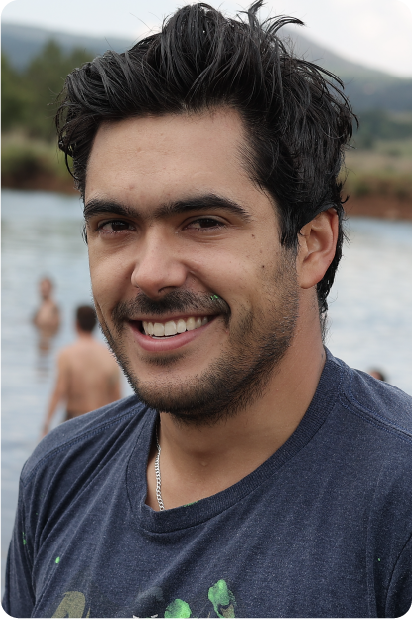 Man with grey beard and curly hair dressed in a dark suit and tie, sitting with fingers interlaced in front of a dark background.