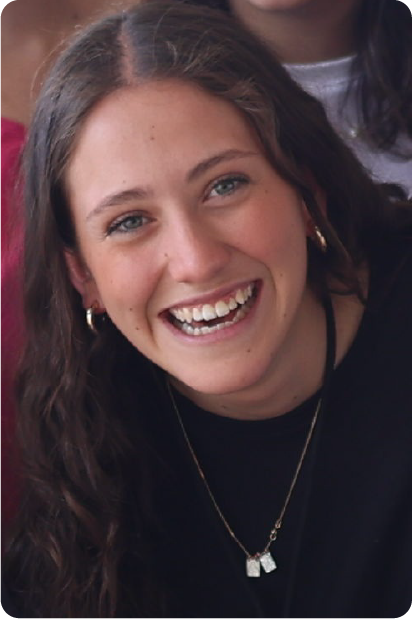 Man with grey beard and curly hair dressed in a dark suit and tie, sitting with fingers interlaced in front of a dark background.