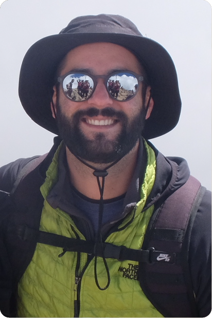 Man with grey beard and curly hair dressed in a dark suit and tie, sitting with fingers interlaced in front of a dark background.