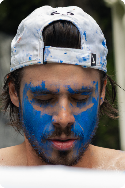 Man with grey beard and curly hair dressed in a dark suit and tie, sitting with fingers interlaced in front of a dark background.