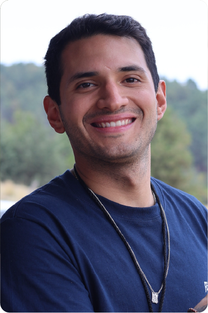 Man with grey beard and curly hair dressed in a dark suit and tie, sitting with fingers interlaced in front of a dark background.