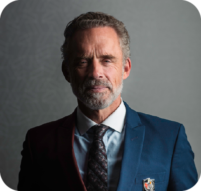 Smiling man with short brown hair wearing a dark jacket and blue shirt outdoors with blurred green foliage background.