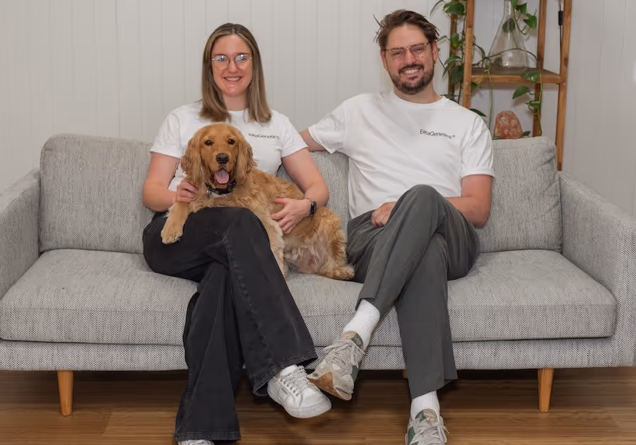A woman and a man sit on a grey couch with a small golden retriever in their lap
