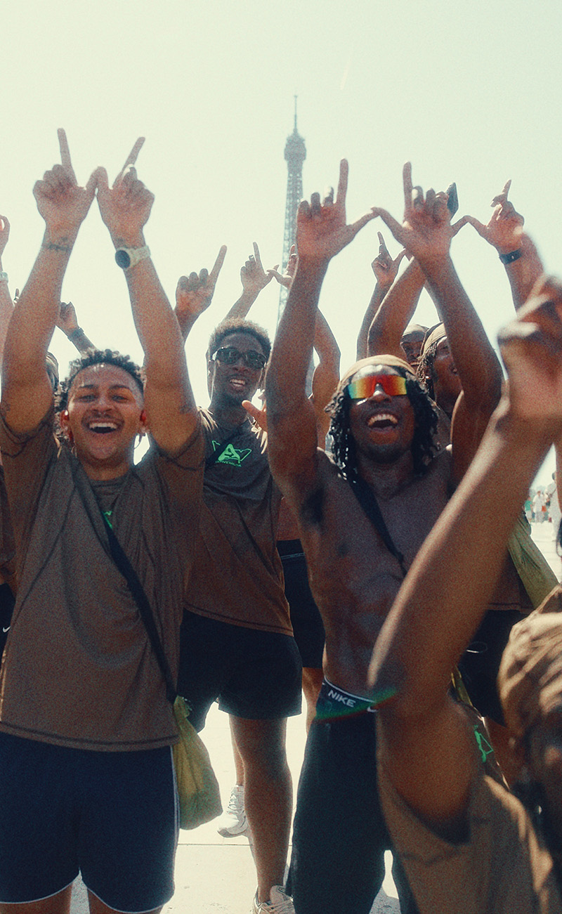 A group of smiling participants in Active Tees raising their hands and making “W” signs in front of the Eiffel Tower, celebrating together in the sun.