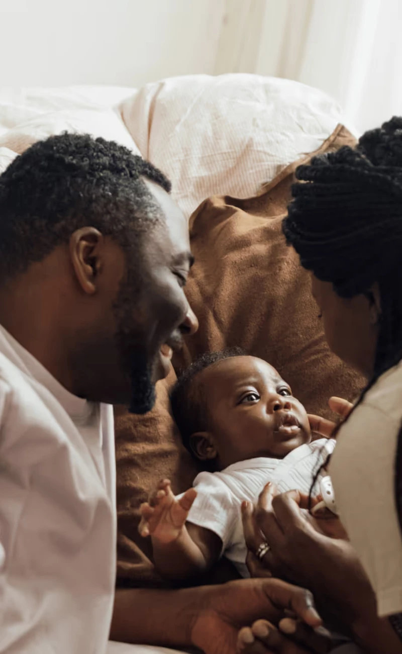 A couple lies on a bed smiling and engaging lovingly with their baby, who looks up at them. The scene feels warm and intimate, with soft natural light and cozy tones.
