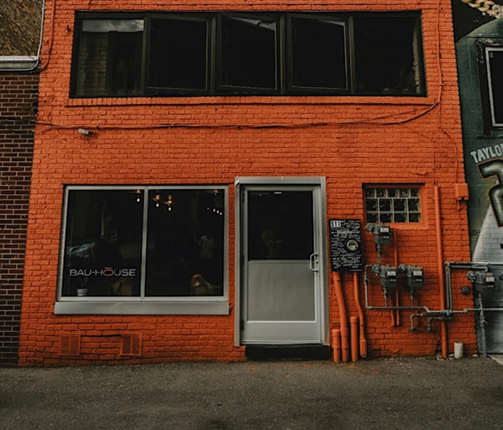 Exterior of Bau-Hōuse Flint, Michigan storefront with orange brick facade, window with logo, gray door, alley view, and utility meters.