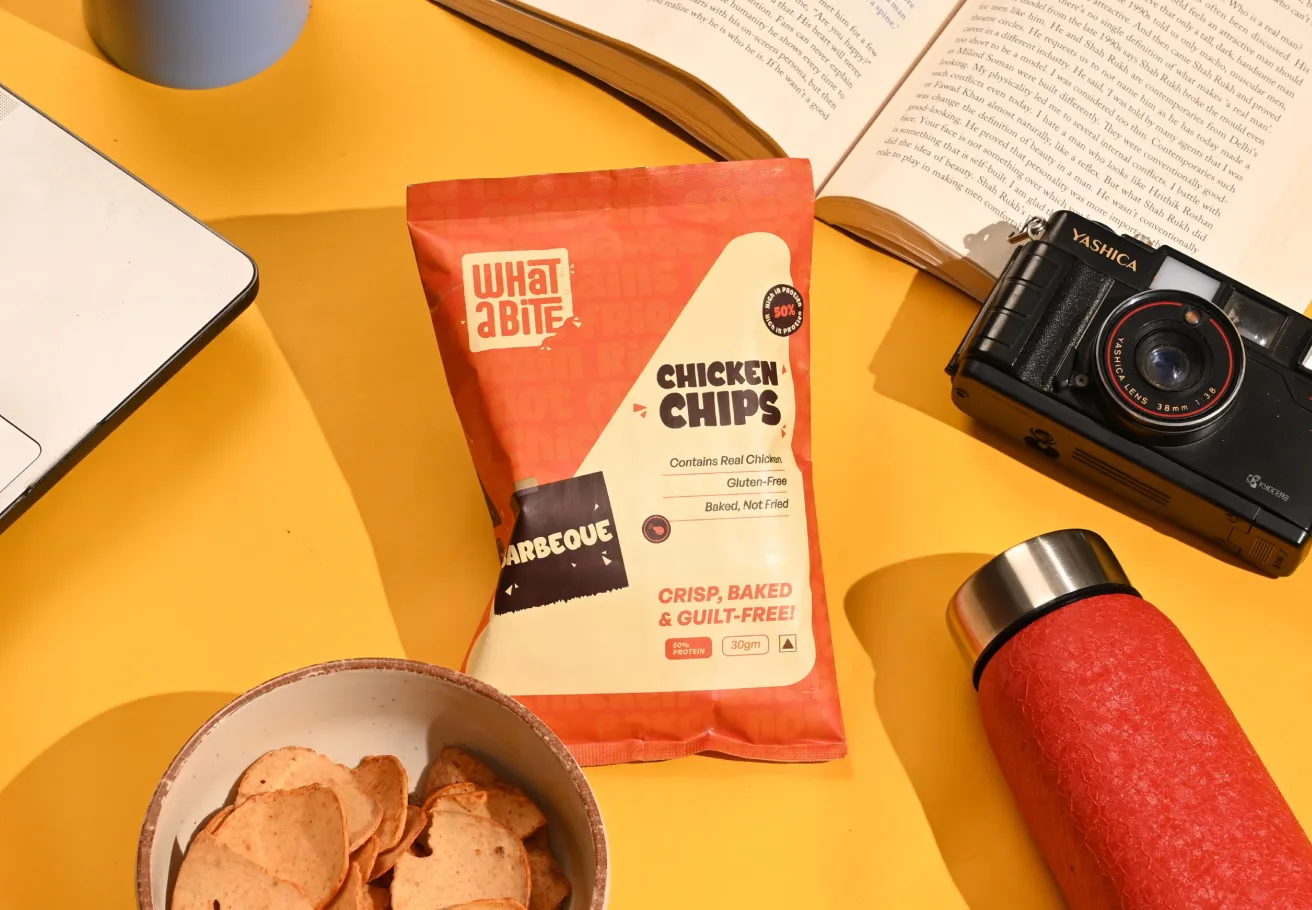 Close-up of a bag of orange-red 'WhatABite Chicken Chips (Barbecue)' resting on a bright yellow surface, surrounded by a laptop, an open book, a black vintage-style camera, a red thermos, and a small white bowl holding some of the chips.