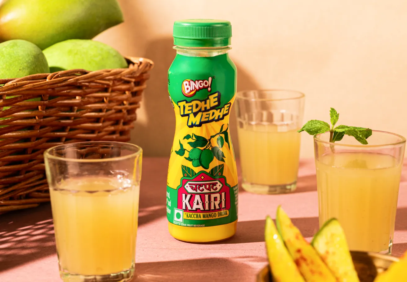 A product photograph showing a green bottle of 'Bingo! Chatpat Kairi' drink, surrounded by glasses of mango juice, a woven basket filled with raw green mangoes, and slices of mango.