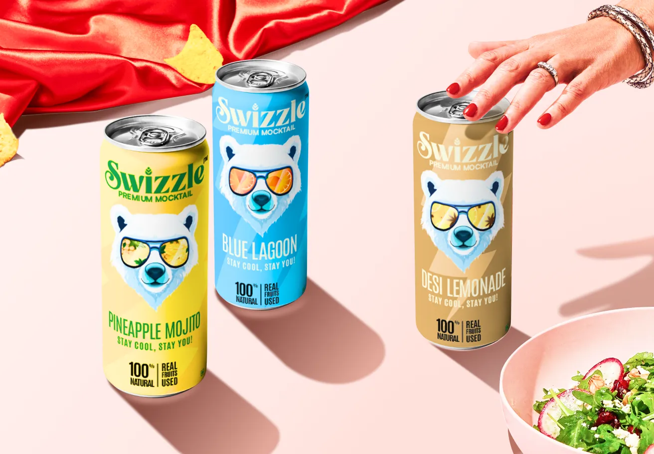 A visually striking product photo featuring three cans of Swizzle Premium Mocktails (Pineapple Mojito, Blue Lagoon, and Desi Lemonade), each bearing a polar bear mascot wearing sunglasses. They are arranged on a pink surface next to a red cloth and a bowl of salad, with a hand reaching for the can on the right.