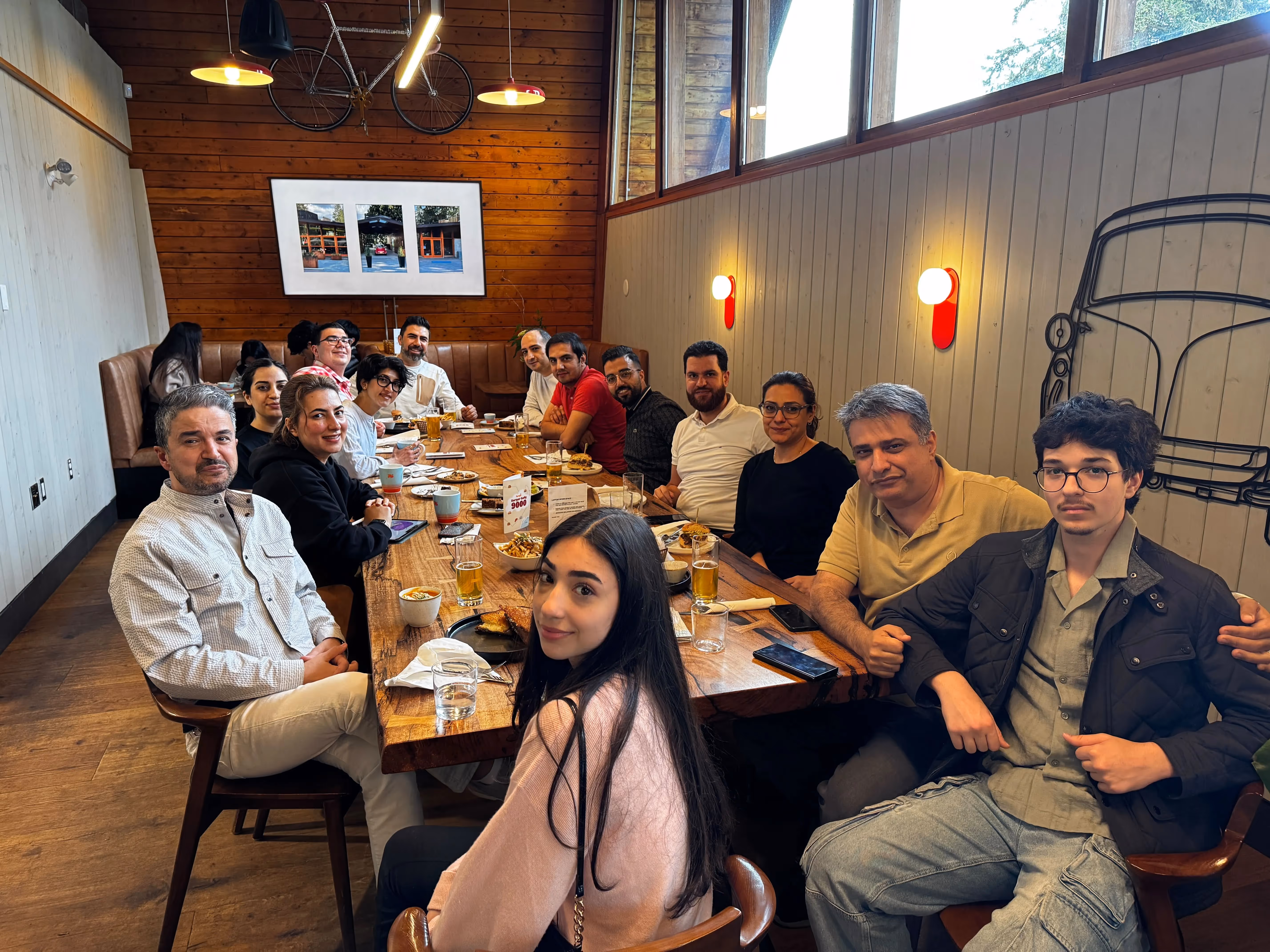 Group of 14 people sitting around a wooden table in a cozy restaurant with drinks and food, smiling at the camera.