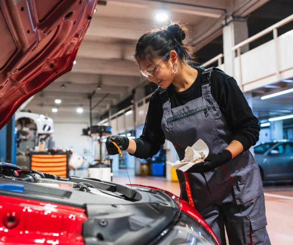 Female mechanic wearing safety glasses and gloves checking engine oil level on a red car in a garage.