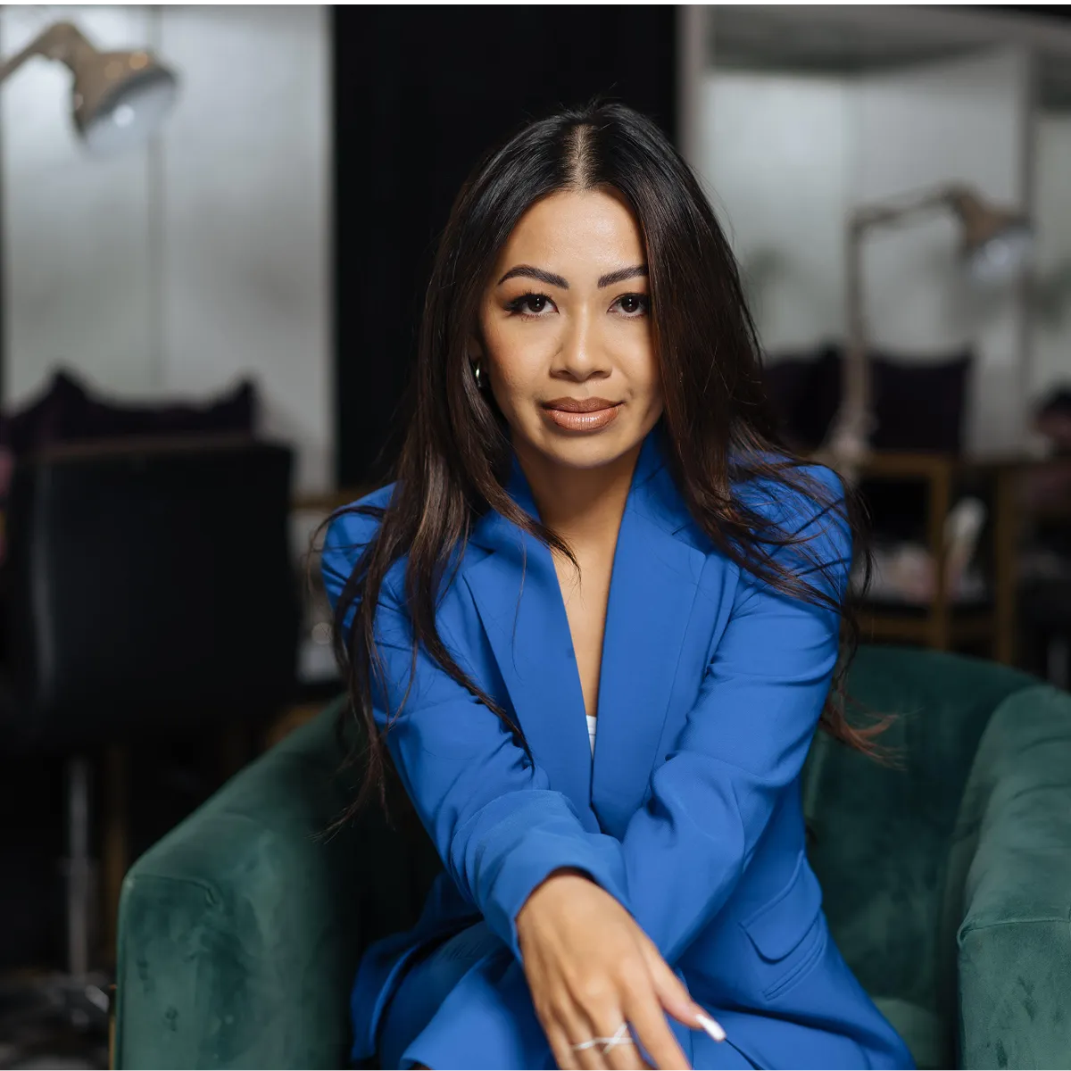 Woman with long dark hair wearing a blue blazer sitting on a green chair, looking confidently at the camera.