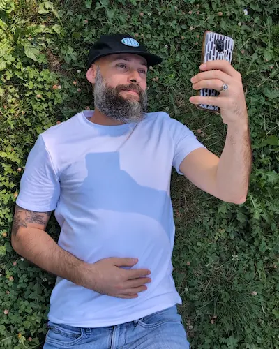 Un homme barbu portant un t-shirt blanc et une casquette noire allongé dans l'herbe et regardant son smartphone. 
