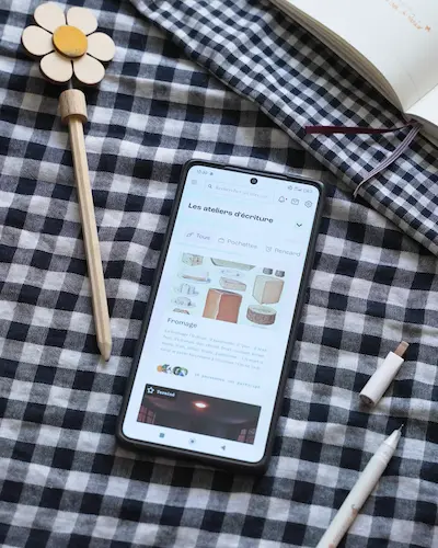 Smartphone ouvert sur pochade la plateforme d'écriture, disposé sur un décor composé d'un tissu motif vichy ainsi qu'un carnet ouvert et un crayon de papier. 